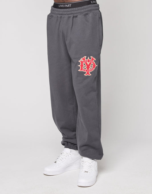 A person wears LIVE FAST DIE YOUNGs DY Jogger in an oversize fit dark gray cotton with a red and white graphic on the upper thigh, white sneakers, and a black elastic waistband featuring the LIVE FAST logo, set against a plain light background.