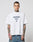 A tattooed man wears the oversize fit LFDY Triple Racing Tee by LIVE FAST DIE YOUNG, a white 100% Baumwolle shirt with LIVE FAST and 40 in blue, paired with blue jeans. He stands against a plain light background, facing the camera.