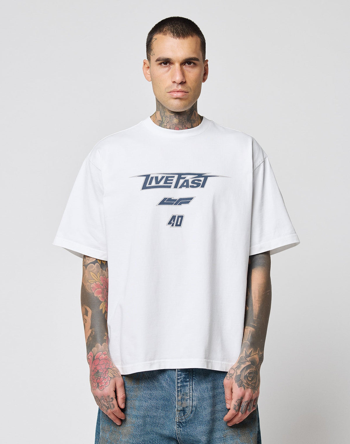 A tattooed man wears the oversize fit LFDY Triple Racing Tee by LIVE FAST DIE YOUNG, a white 100% Baumwolle shirt with LIVE FAST and 40 in blue, paired with blue jeans. He stands against a plain light background, facing the camera. | model