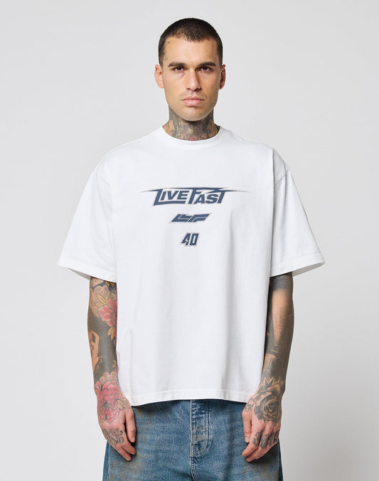 A tattooed man wears the oversize fit LFDY Triple Racing Tee by LIVE FAST DIE YOUNG, a white 100% Baumwolle shirt with LIVE FAST and 40 in blue, paired with blue jeans. He stands against a plain light background, facing the camera. | model