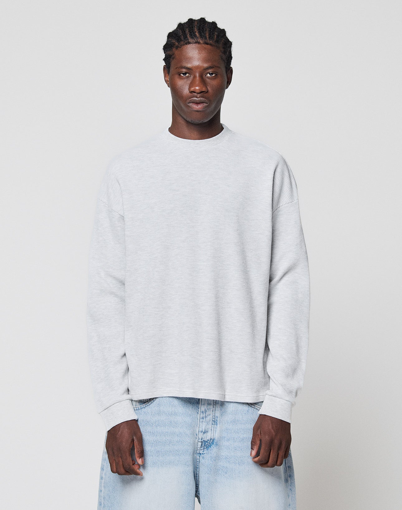 A man with braided hair wears the LIVE FAST DIE YOUNG Blank Waffle Longsleeve in light gray and light blue jeans, standing against a plain white background and looking straight ahead with a neutral expression. | model