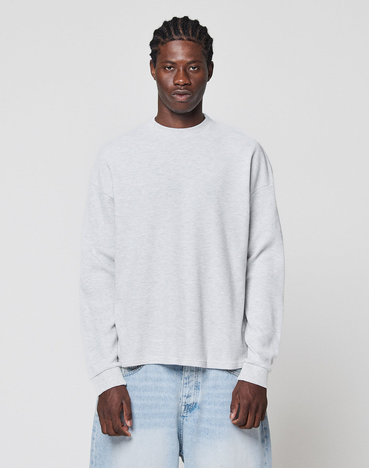 A man with braided hair wears the LIVE FAST DIE YOUNG Blank Waffle Longsleeve in light gray and light blue jeans, standing against a plain white background and looking straight ahead with a neutral expression. | model