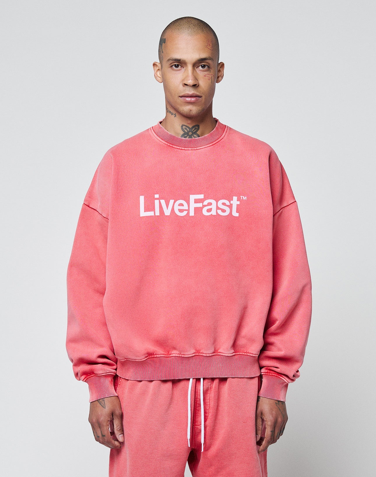 A person wears the oversize Basic LiveFast Sweater by LIVE FAST DIE YOUNG in faded pink, featuring a bold frontprint and paired with matching sweatpants, standing against a plain light gray background.