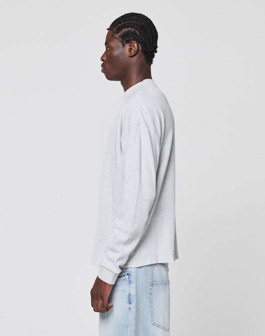 A person with braided hair stands in profile, wearing the LIVE FAST DIE YOUNG Blank Waffle Longsleeve in light gray and light blue jeans, against a plain white background.