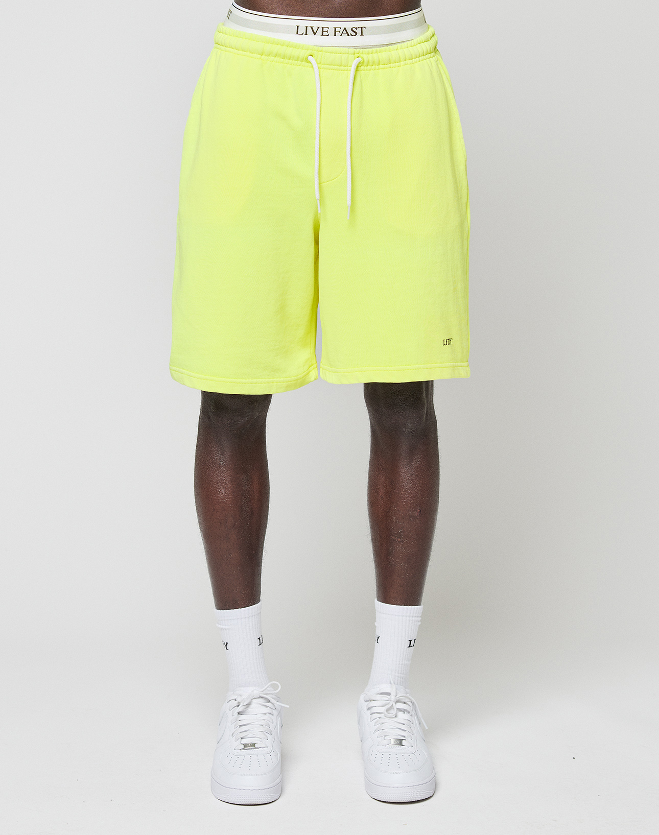 A person wearing LIVE FAST DIE YOUNG Basic Shorts in bright yellow with a logo print and elastic waistband, plus white socks and sneakers, stands against a plain white background. The shorts’ waistband displays the LIVE FAST text. Only the lower half is shown.