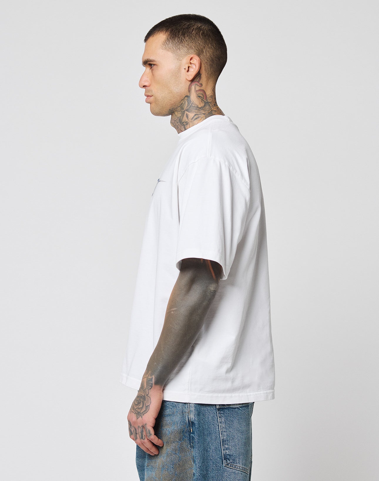 A man with short hair and tattoos on his neck and arm stands in profile wearing the LFDY Triple Racing Tee by LIVE FAST DIE YOUNG—an oversize fit white T-shirt with multilogo frontprint, made from 100% Baumwolle, paired with blue jeans against a gray background.