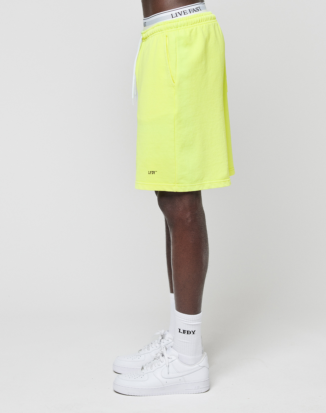 Side view of a person wearing LIVE FAST DIE YOUNG's Basic Shorts in bright yellow with an elastic waistband and logo print, paired with a white LIVE FAST waistband, LFDY socks, and white sneakers against a plain white background.