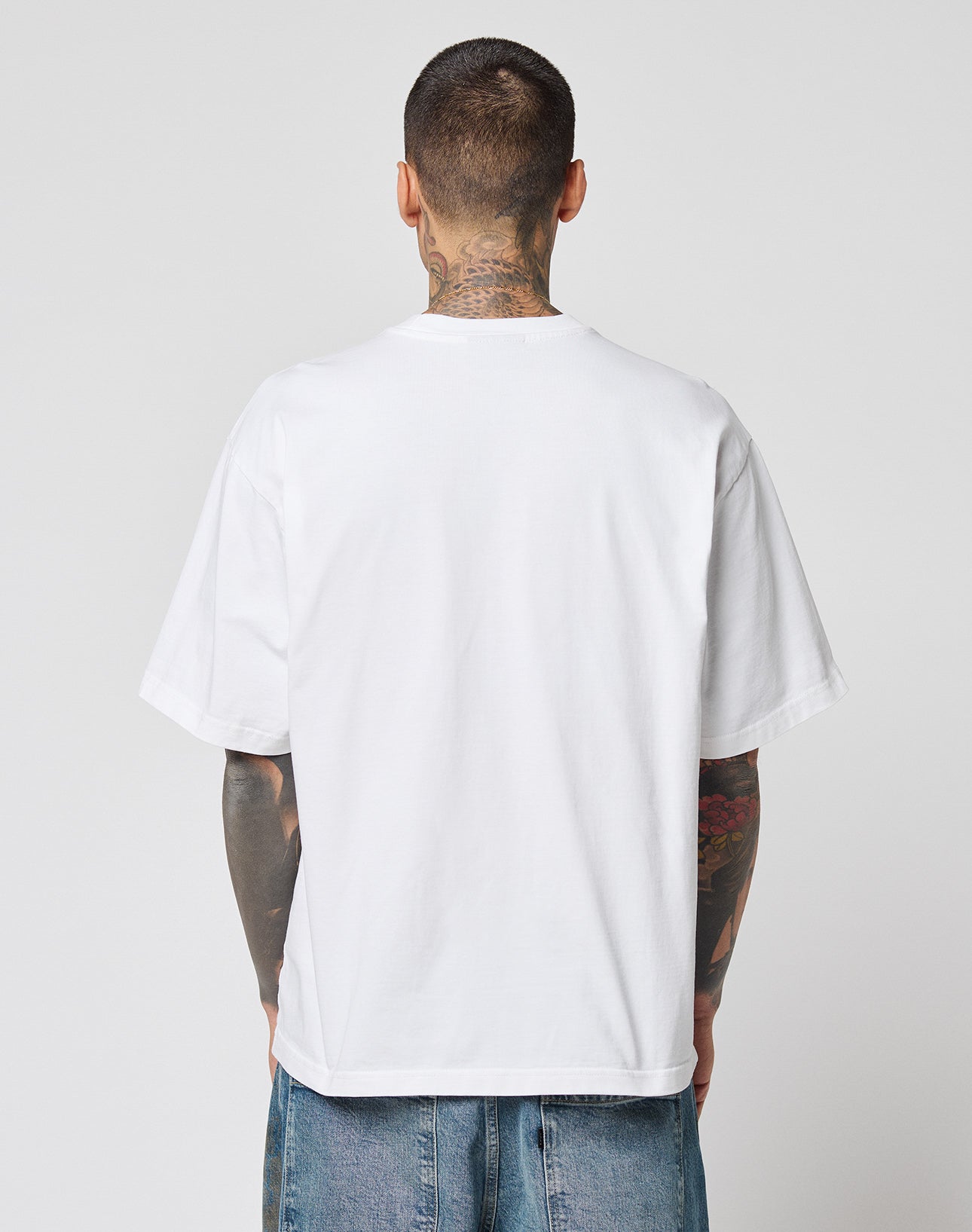 A tattooed man stands with his back to the camera, wearing a white LFDY Triple Racing Tee by LIVE FAST DIE YOUNG and blue jeans, set against a light gray background.