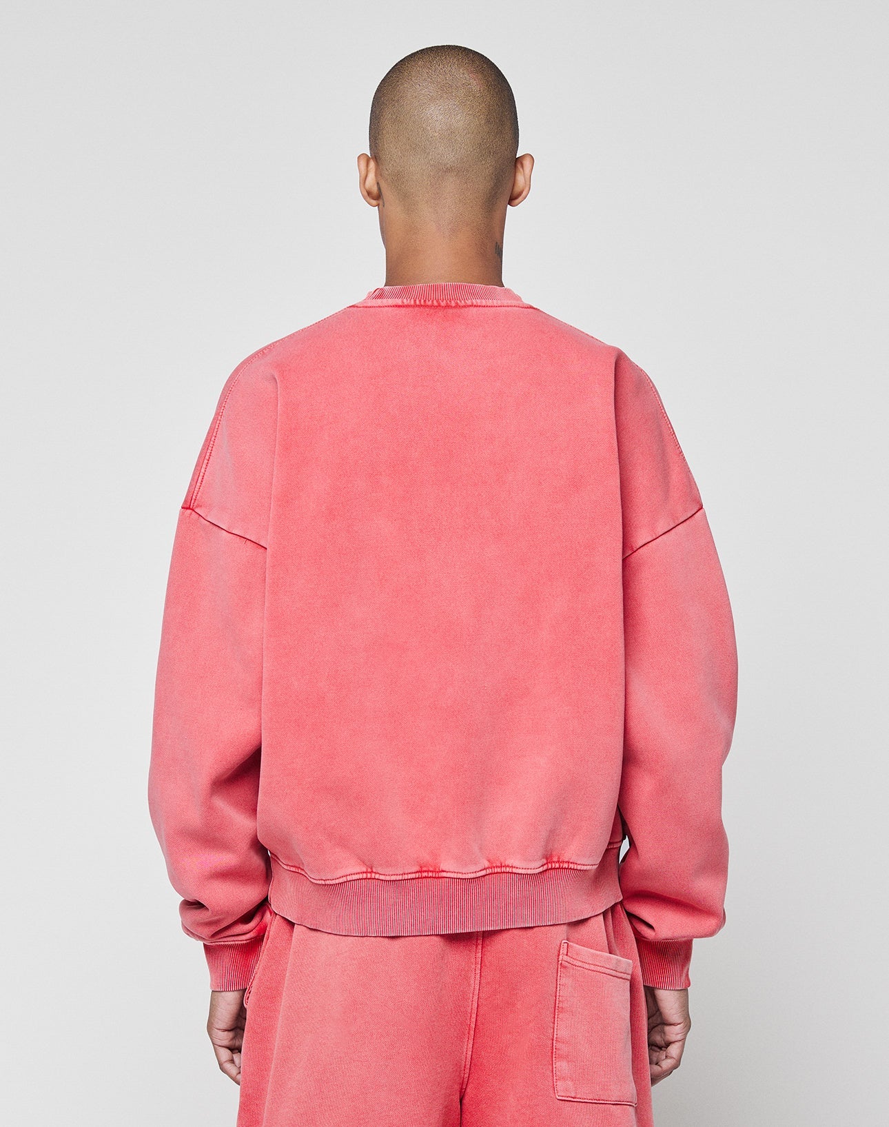 A person with a shaved head faces away from the camera, wearing the oversize Basic LiveFast Sweater and matching sweatpants by LIVE FAST DIE YOUNG in faded pink, set against a plain light gray background.