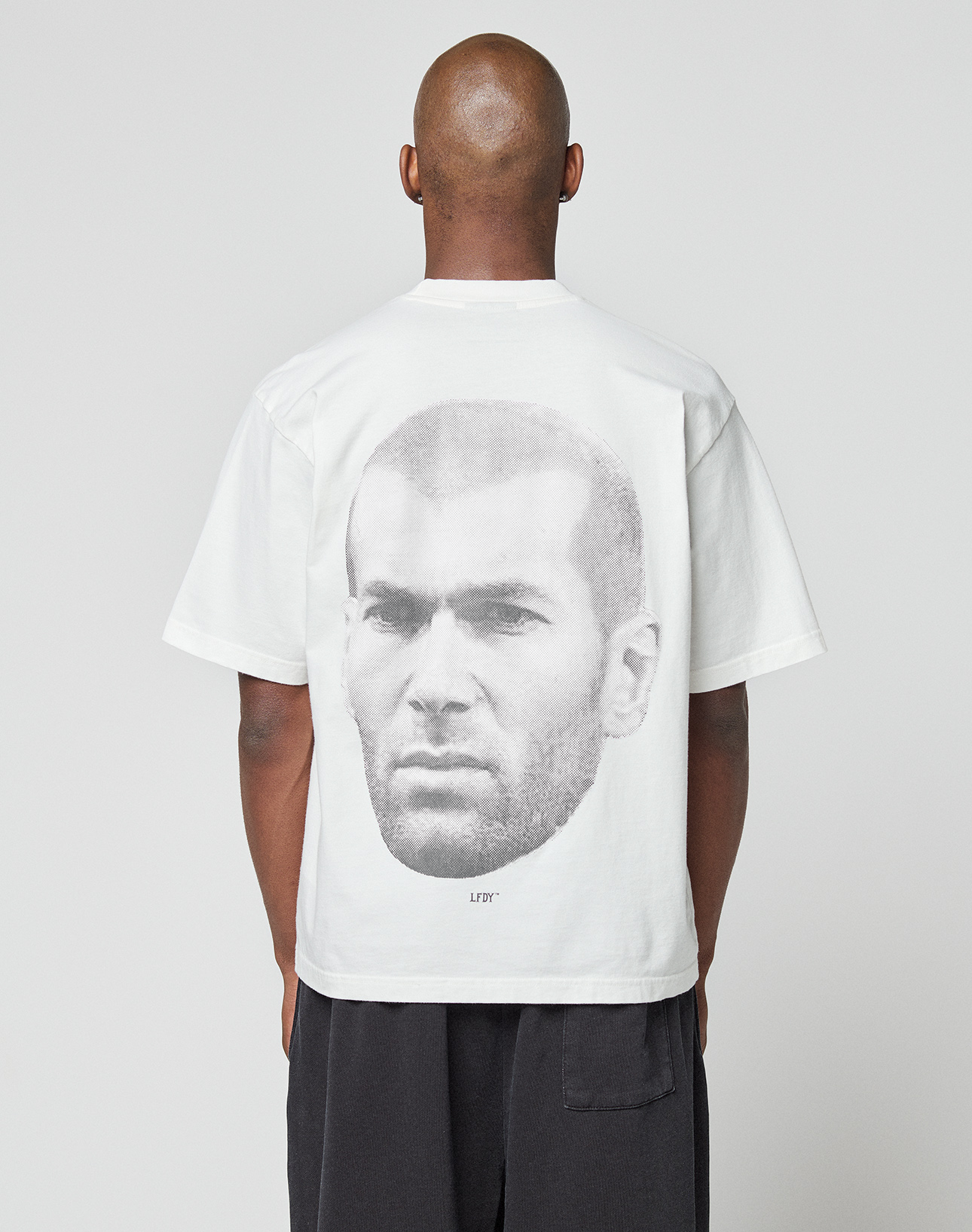 A person with a shaved head wears the LIVE FAST DIE YOUNG LFDY Zizou Tee, an oversized white 100% Baumwolle t-shirt featuring short sleeves and a bold black-and-white portrait of a man's face as the backprint. | model