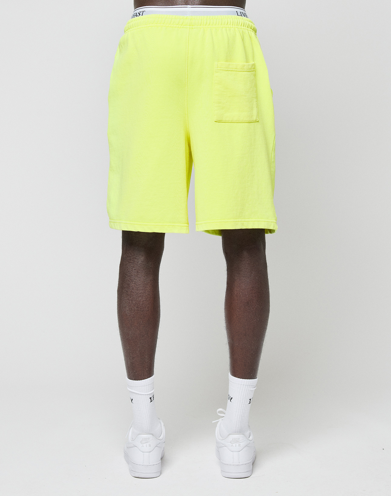 A person with their back to the camera wears LIVE FAST DIE YOUNG Basic Shorts in bright yellow, featuring a regular fit, elastic waistband, and back pocket, paired with white socks and sneakers against a plain light background.