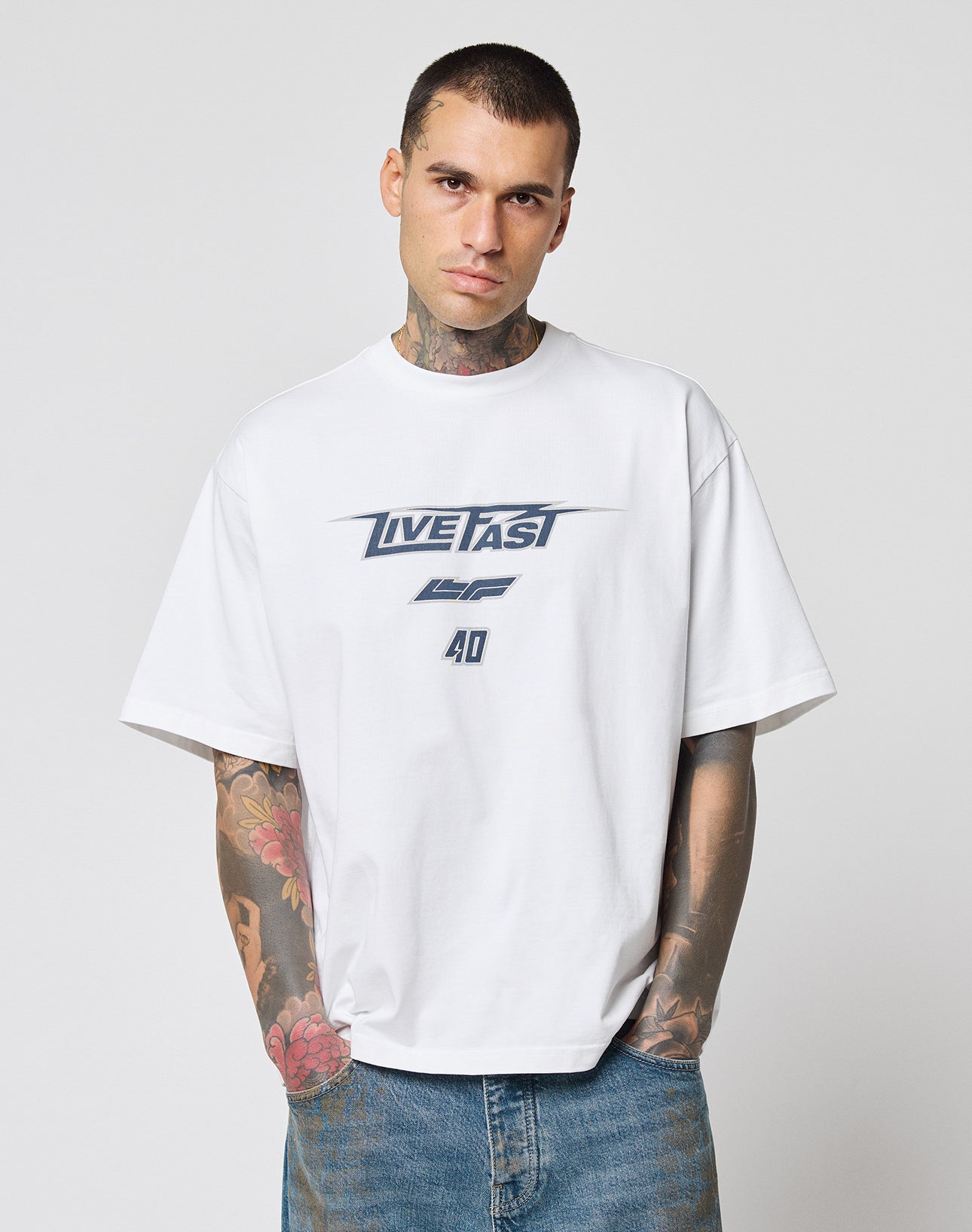 A man with short hair and visible arm tattoos wears the LIVE FAST DIE YOUNG LFDY Triple Racing Tee in white and blue jeans, standing against a plain light background and looking directly at the camera.