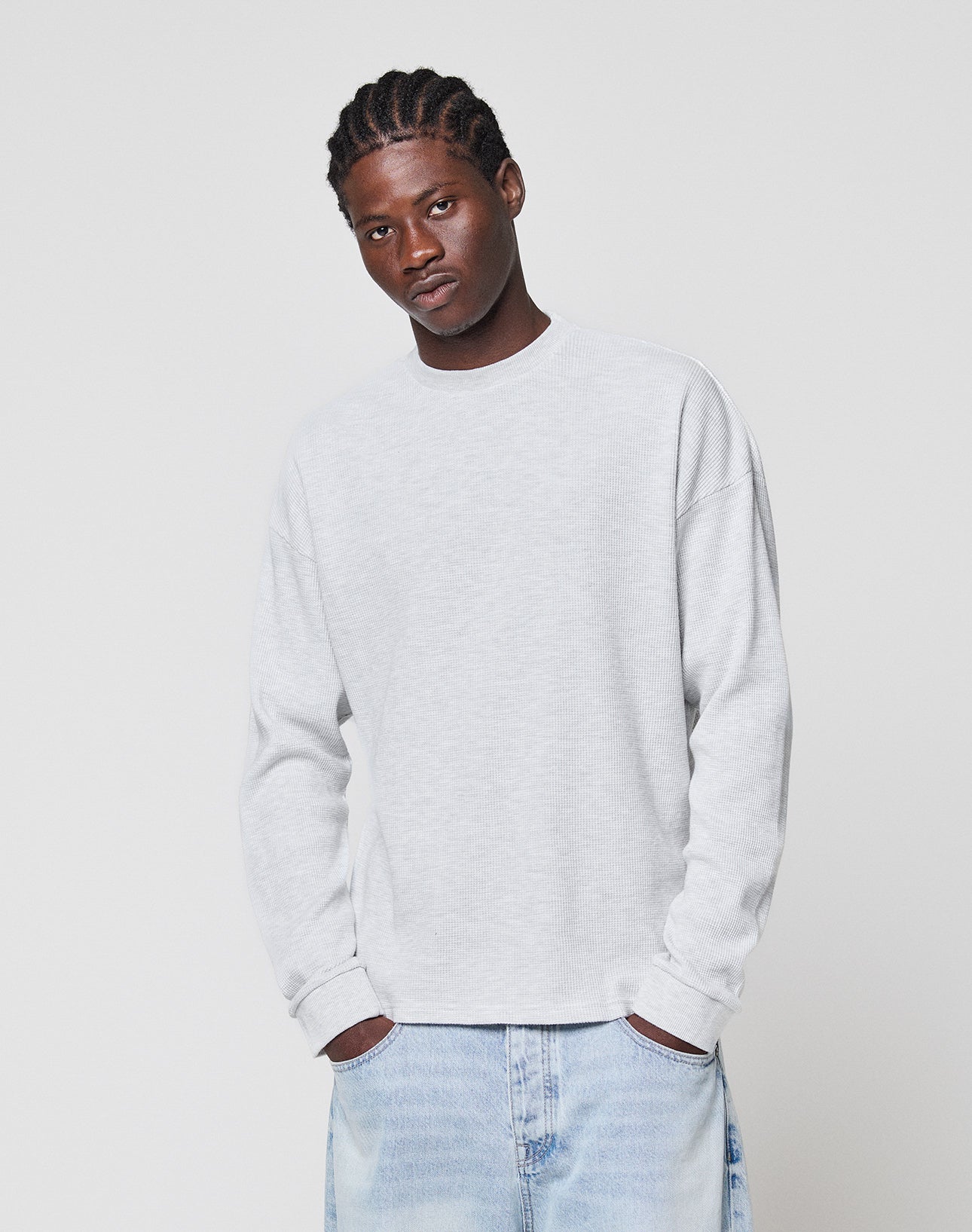 A man with braided hair wears the LIVE FAST DIE YOUNG Blank Waffle Longsleeve—an oversize fit, light gray 100% Baumwolle sweatshirt—with light blue jeans, standing against a plain white background, expression neutral.