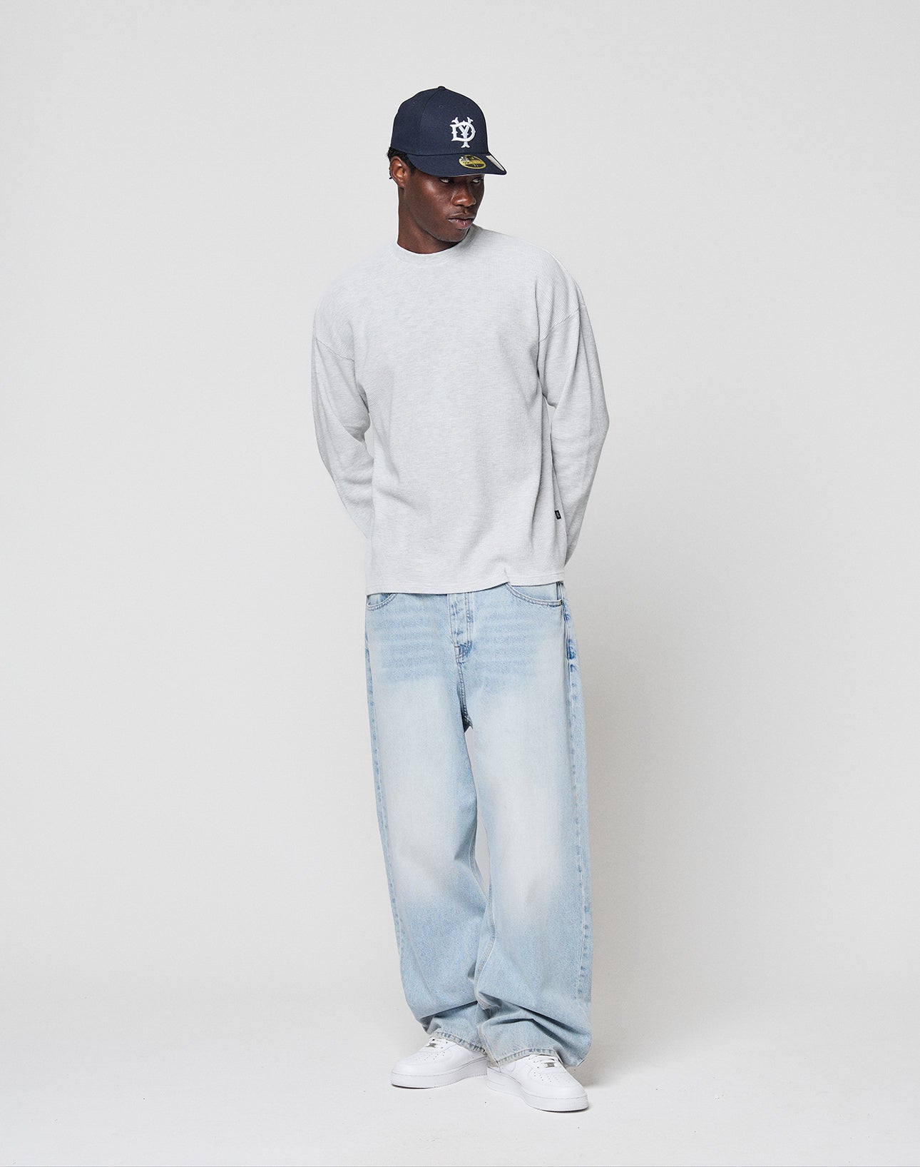 A man wears the LIVE FAST DIE YOUNG Blank Waffle Longsleeve in light gray, a navy baseball cap, baggy light blue jeans, and white sneakers. He stands against a plain light gray background with hands behind his back, looking left.