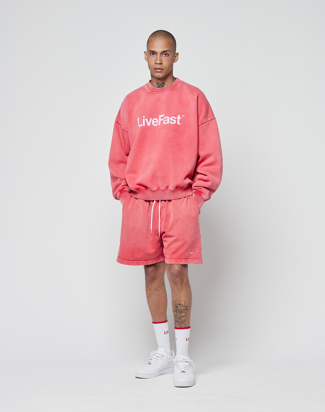 A person with short hair and visible neck and leg tattoos wears a pink Basic LiveFast Sweater by LIVE FAST DIE YOUNG, matching pink shorts, white crew socks, and white sneakers, standing against a plain background.