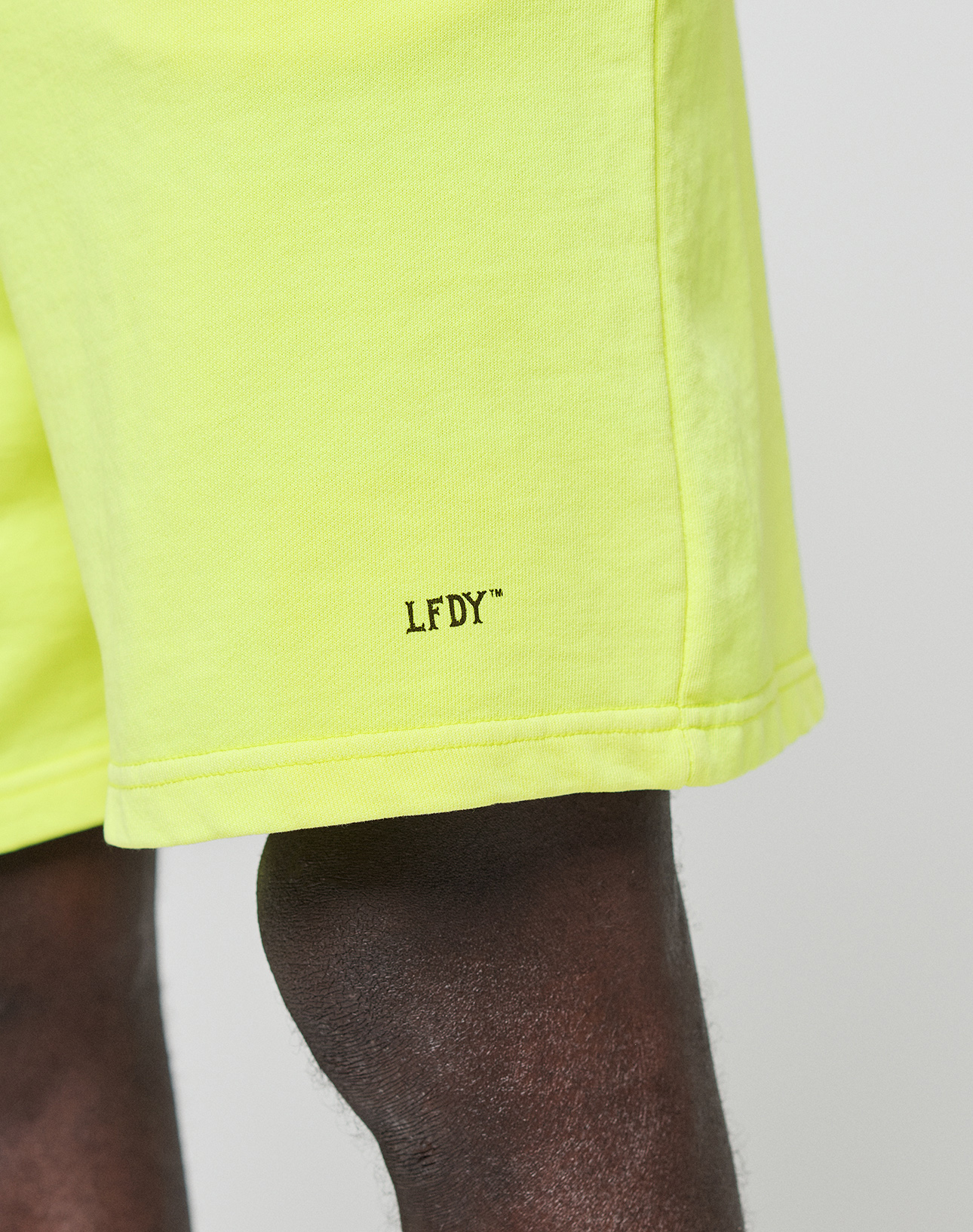 Close-up of a person wearing LIVE FAST DIE YOUNG Basic Shorts in bright yellow with "LFDY™" logo near the hem. These regular fit shorts have an elastic waistband, shown against a plain background that highlights the wearer's dark skin.