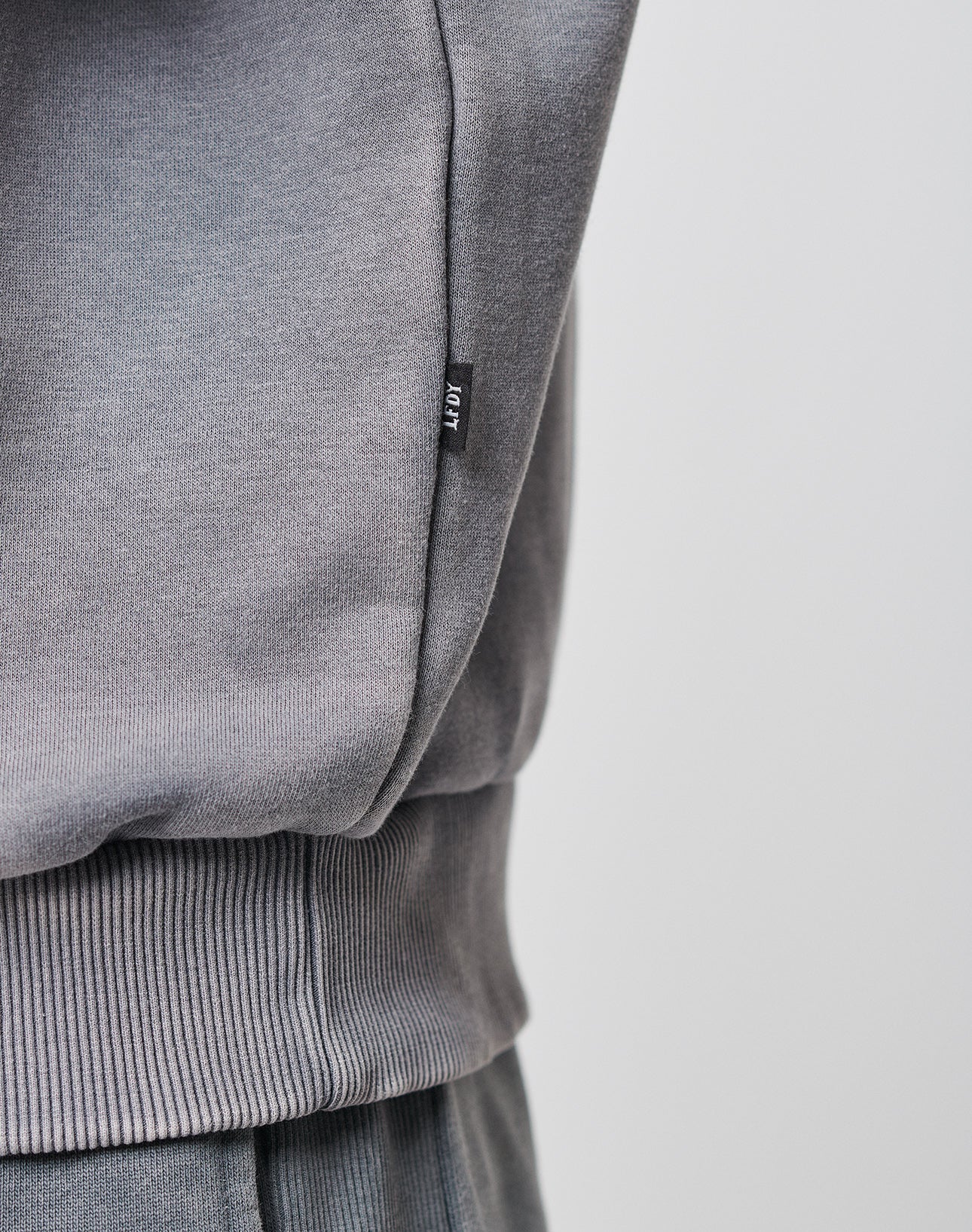 Close-up of a person wearing the Basic Blank Hooded and matching pants by LIVE FAST DIE YOUNG in grey ribbed fabric, Oversize Fit. A small black tag with white text is on the side seam. The background is plain and light-colored.