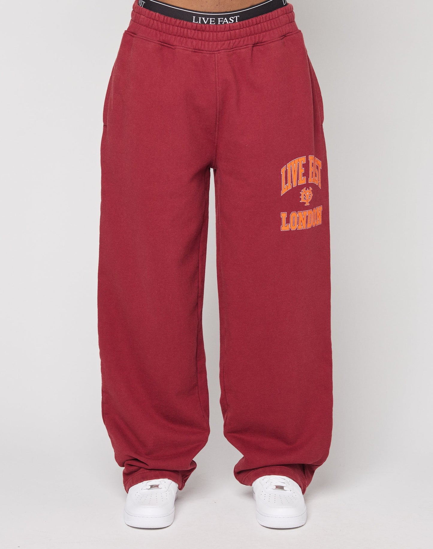 Person wearing LIVE FAST DIE YOUNGs Live Fast London Wide Jogger in maroon with a yellow lion emblem and LIVE FAST LONDON text on the left leg, white sneakers, and a matching elastic waistband with the LIVE FAST logo.