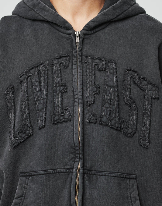 Close-up of a person wearing the Ripped College Hooded Zip by LIVE FAST DIE YOUNG, an oversized black zip-up hoodie with LIVE FAST embroidered in large letters across the chest. The persons face is not shown.
