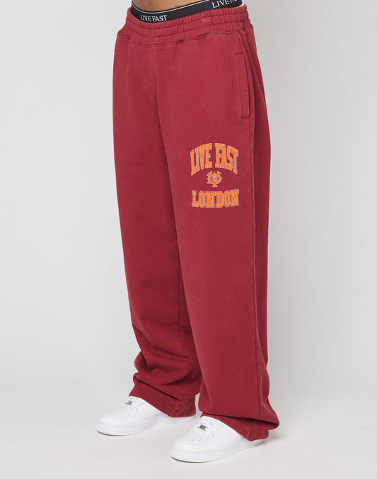 Lower body view of a person in LIVE FAST DIE YOUNG’s Live Fast London Wide Jogger—loose-fitting red sweatpants featuring “LIVE FAST LONDON” and a yellow lion graphic on the left leg—paired with white sneakers. | model