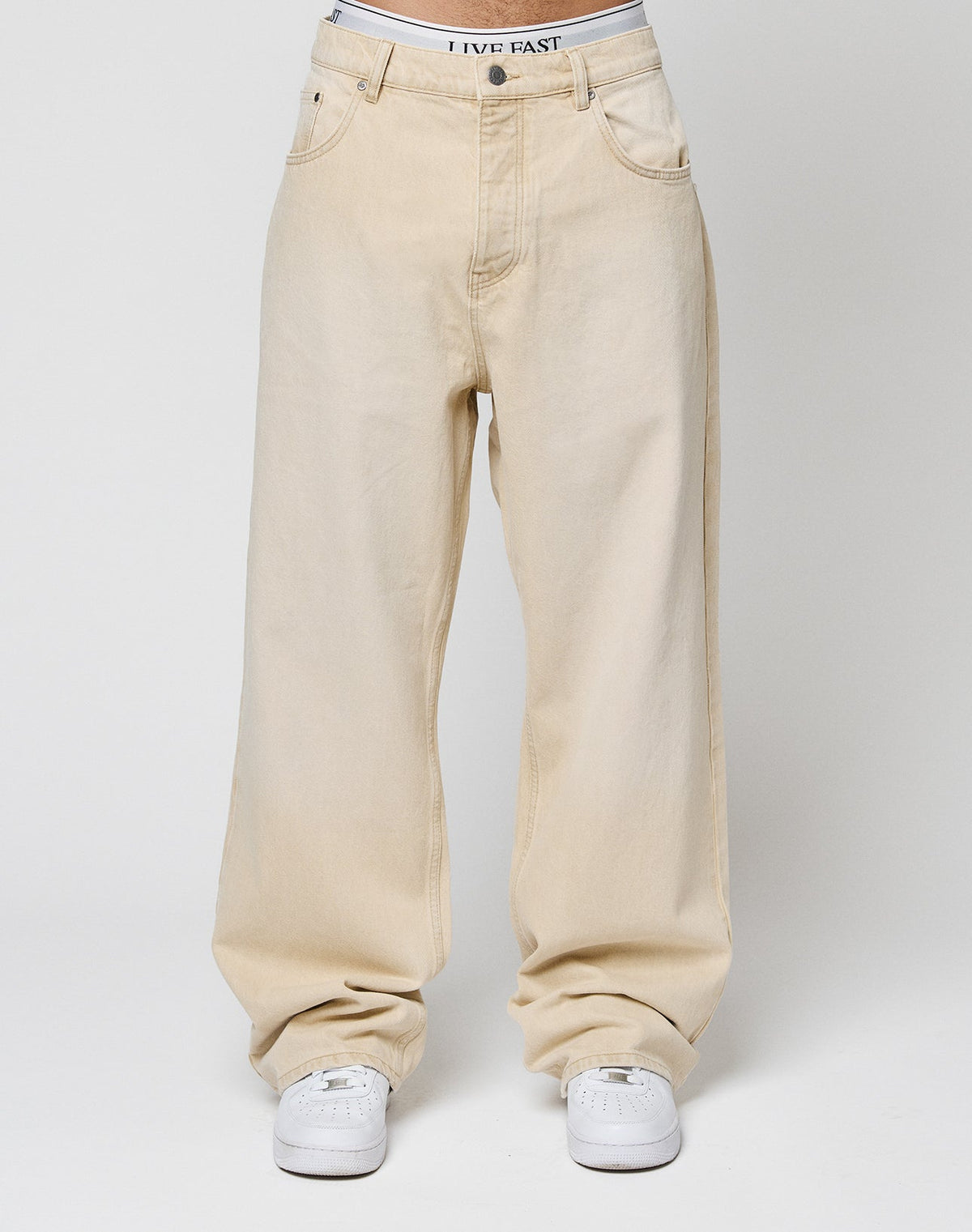 Someone wearing the LIVE FAST DIE YOUNG LFDY Baggy Denim in light beige—100% Baumwolle, baggy fit with weites gerades Bein and a LIVE FAST waistband—paired with white sneakers, stands against a plain white background.