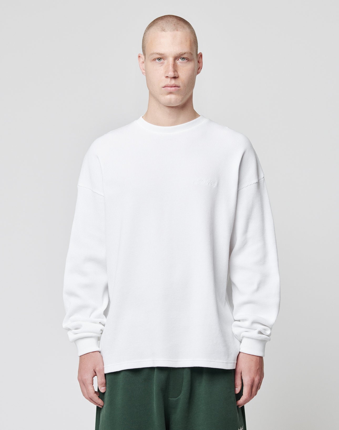 A person with a shaved head wears the LIVE FAST DIE YOUNG Basic Script Waffle Longsleeve in white and dark green pants, standing against a light gray background with a neutral expression.