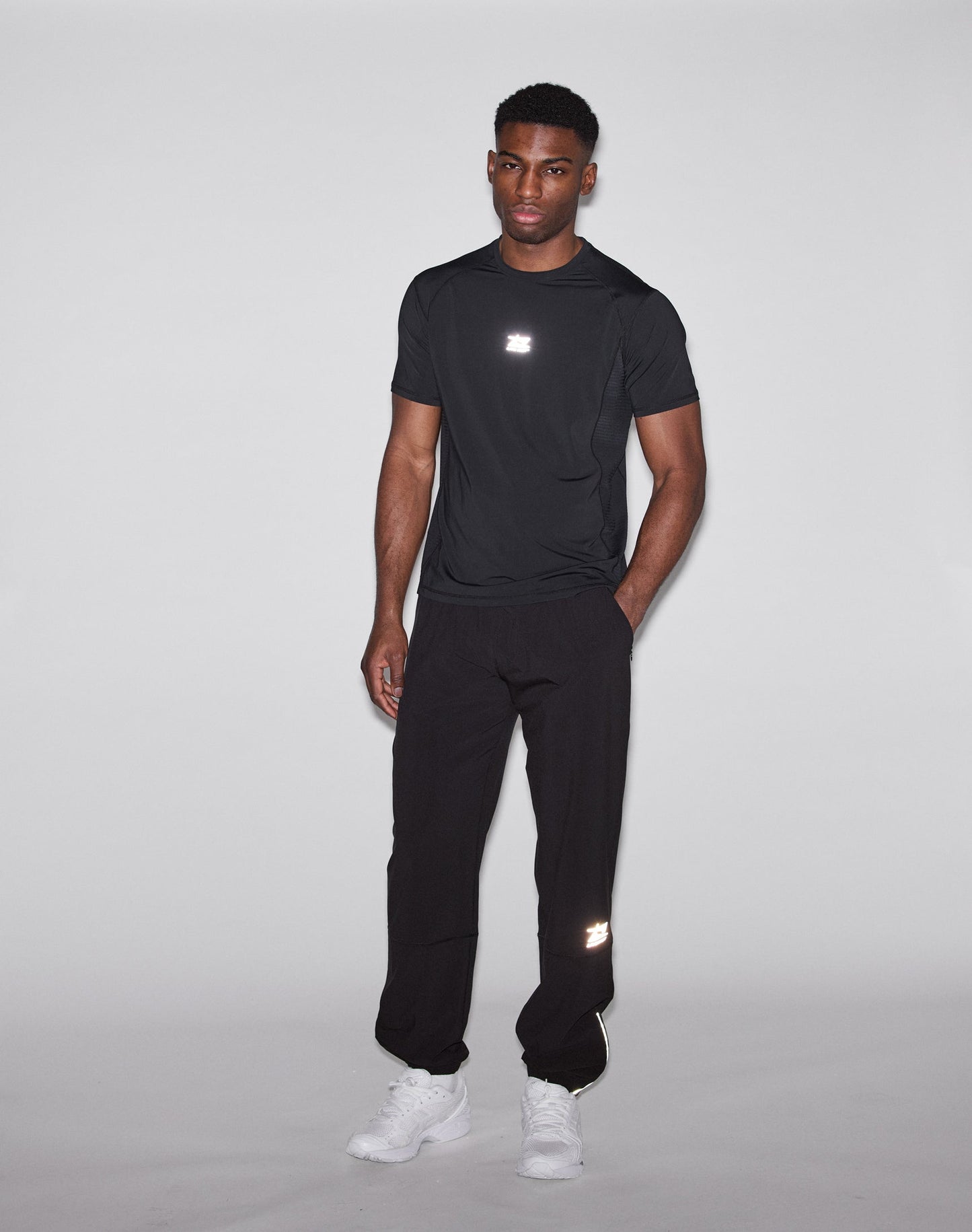 A man faces the camera against a light background in the LIVE FAST DIE YOUNG Live Fast Performance Tee, slim fit, black with reflective front and back print, black pants, and white sneakers. His arms rest casually at his sides.