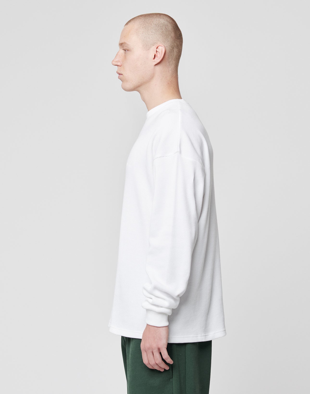 A person with a shaved head stands in profile, facing left, wearing the LIVE FAST DIE YOUNG Basic Script Waffle Longsleeve in white and green pants against a plain light grey background.