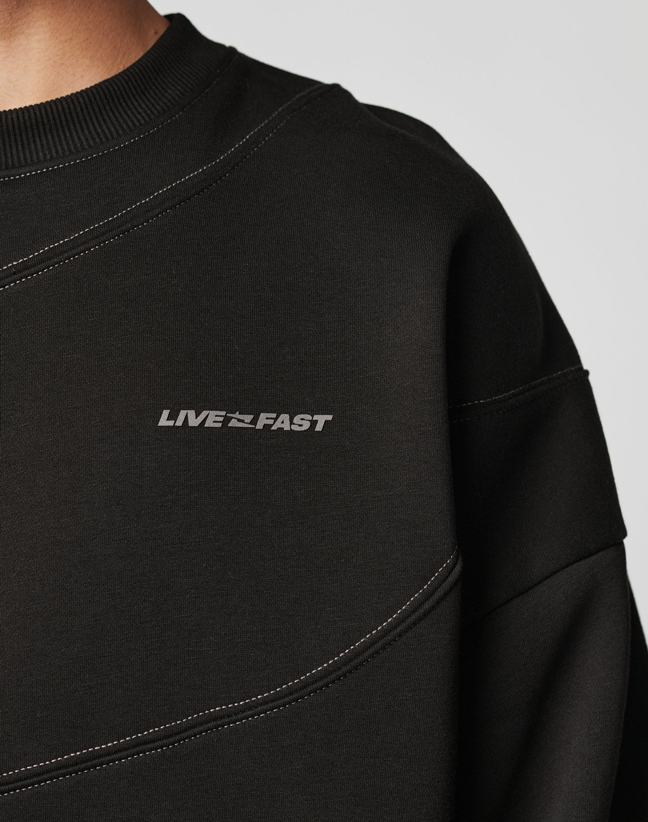 Close-up of a person wearing the LFDY Wavy Sweater by LIVE FAST DIE YOUNG, featuring black Baumwolle fabric, white stitching, and LIVE FAST printed in white on the chest; only shoulder and part of neck are visible.