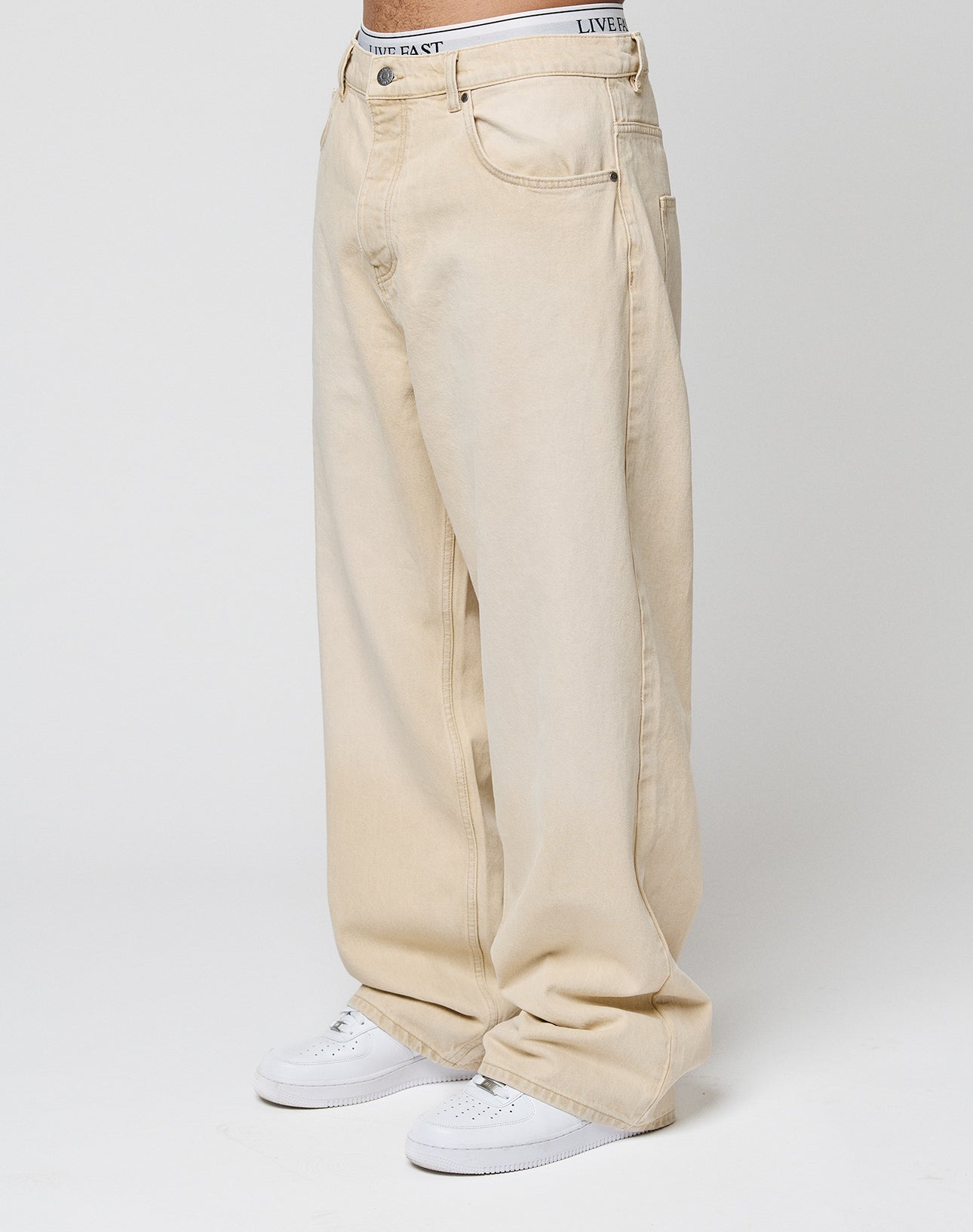 Eine Person trägt LFDY Baggy Denim von LIVE FAST DIE YOUNG aus 100% Baumwolle mit weitem, geradem Bein sowie weißen Sneakern. Vor weißem Hintergrund sind nur Unterkörper und Beine zu sehen.