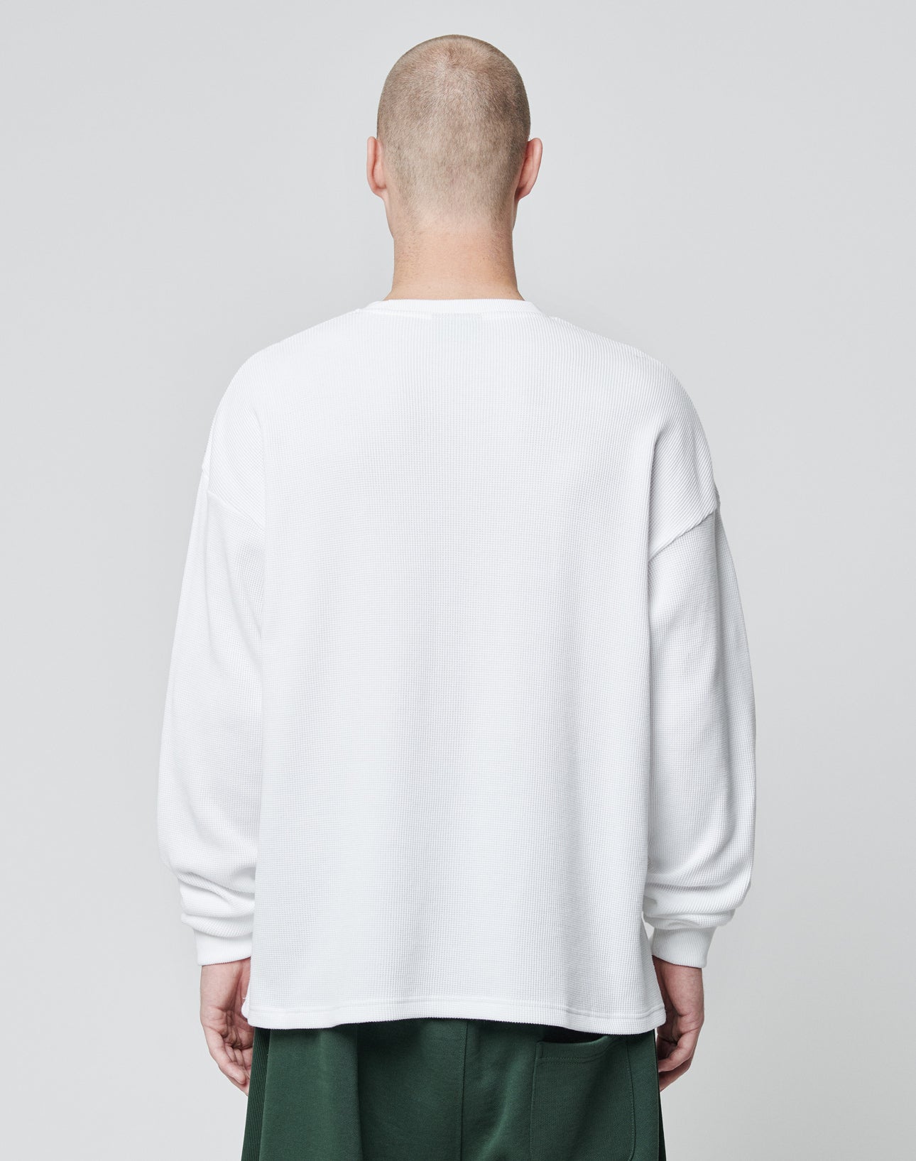 A person with a shaved head wears the LIVE FAST DIE YOUNG Basic Script Waffle Longsleeve, an oversize white 100% Baumwolle shirt, and green pants, standing with their back to a plain light gray background.