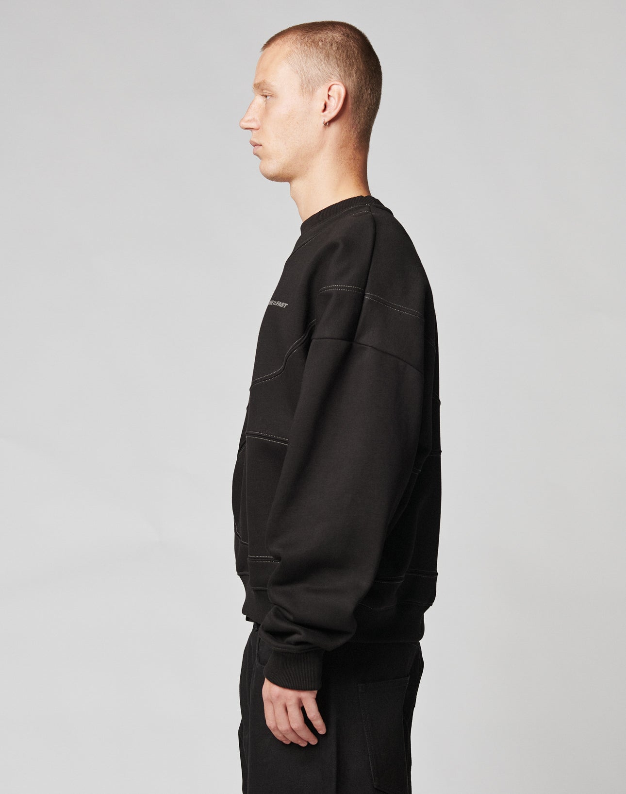 A person with short hair stands in profile facing left, wearing a black LIVE FAST DIE YOUNG LFDY Wavy Sweater and black pants against a plain, light gray background.