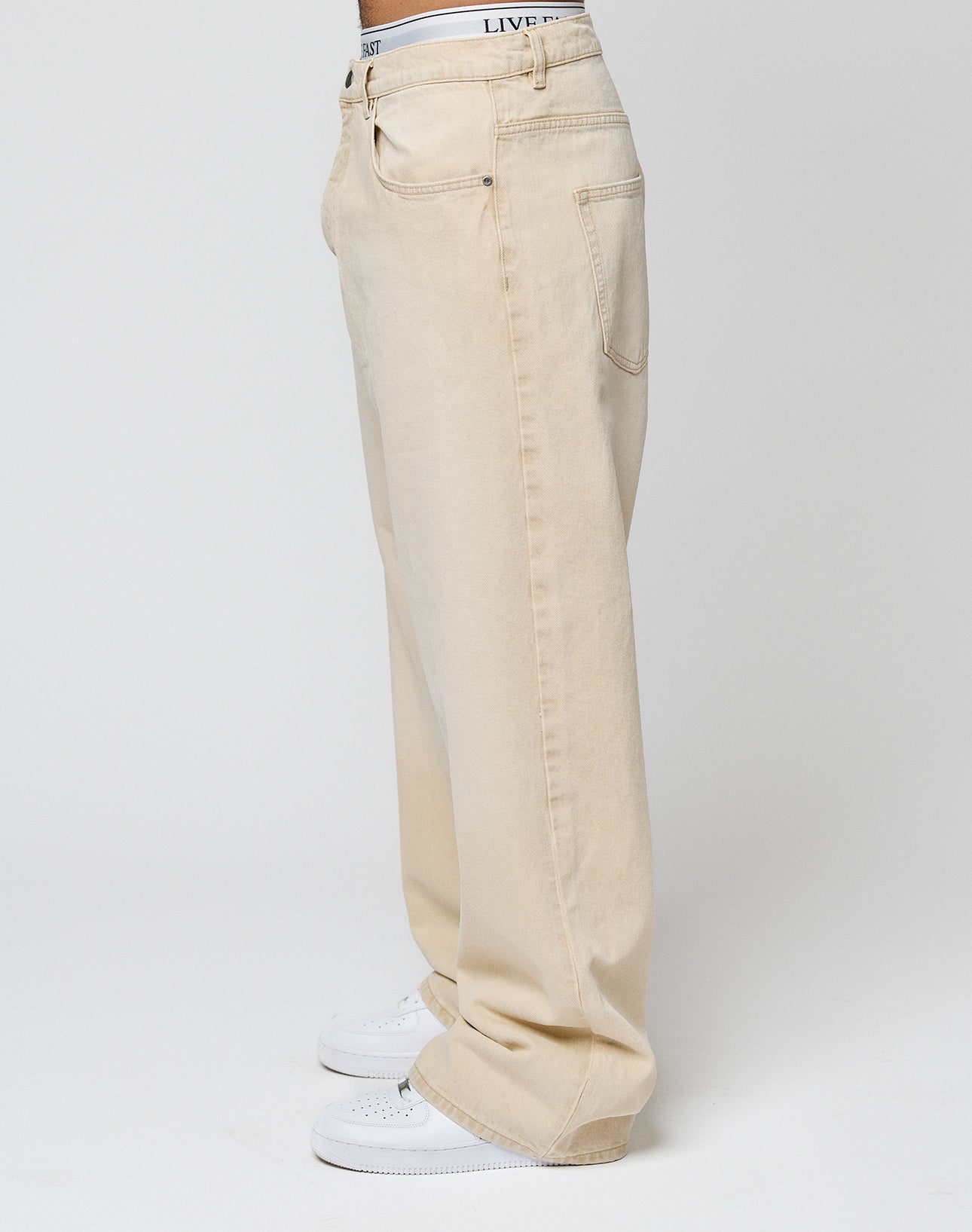 Person steht seitlich und trägt die LFDY Baggy Denim von LIVE FAST DIE YOUNG in hellbeige mit weitem, geradem Bein, sichtbaren Taschen und weißen Sneakern. Der Bund weißer Unterwäsche ist leicht über der Jeans sichtbar. Heller, schlichter Hintergrund.