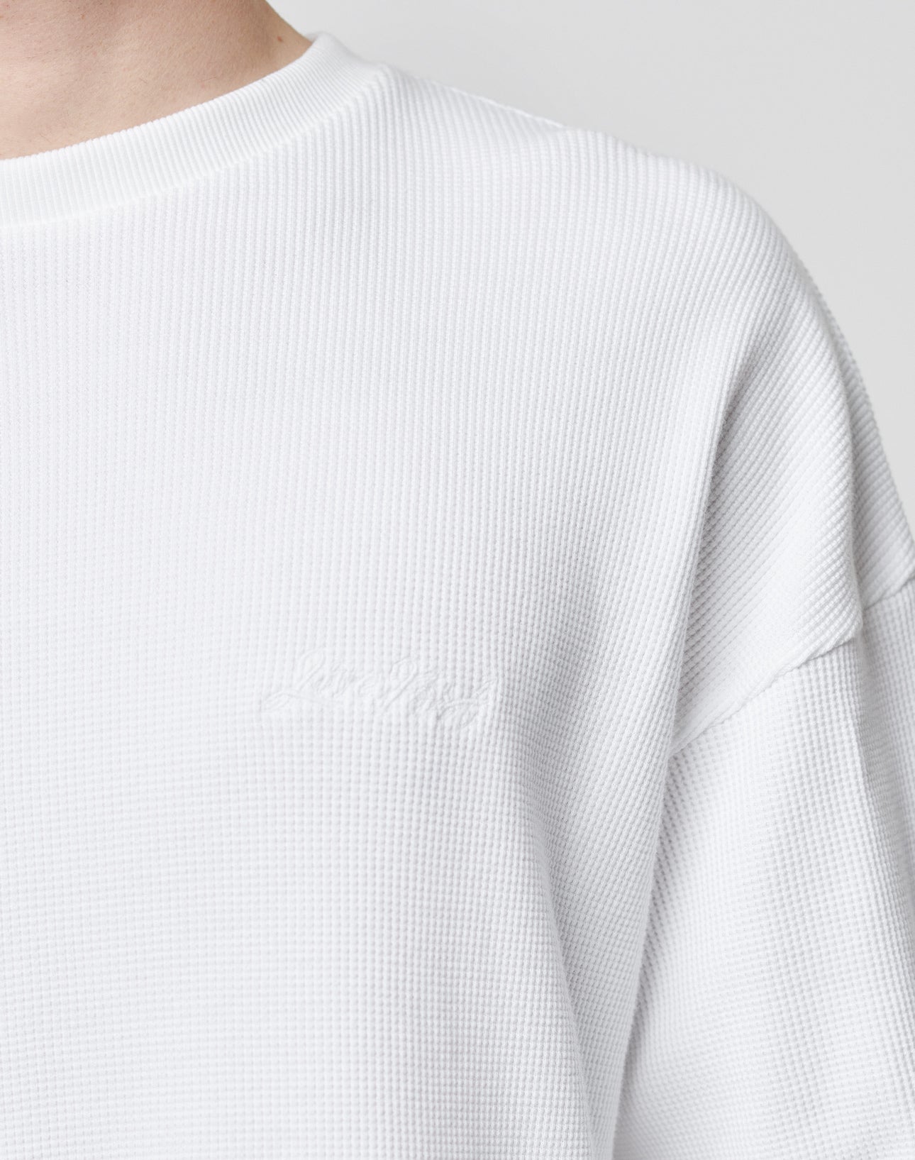 Close-up of a person wearing the LIVE FAST DIE YOUNG Basic Script Waffle Longsleeve, a textured white crew-neck shirt with an oversize fit and subtle logo embroidery near the chest. The background is plain and light-colored.