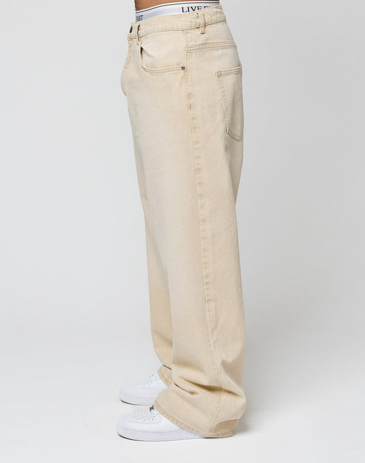 Person steht seitlich und trägt die LFDY Baggy Denim von LIVE FAST DIE YOUNG in hellbeige mit weitem, geradem Bein, sichtbaren Taschen und weißen Sneakern. Der Bund weißer Unterwäsche ist leicht über der Jeans sichtbar. Heller, schlichter Hintergrund.