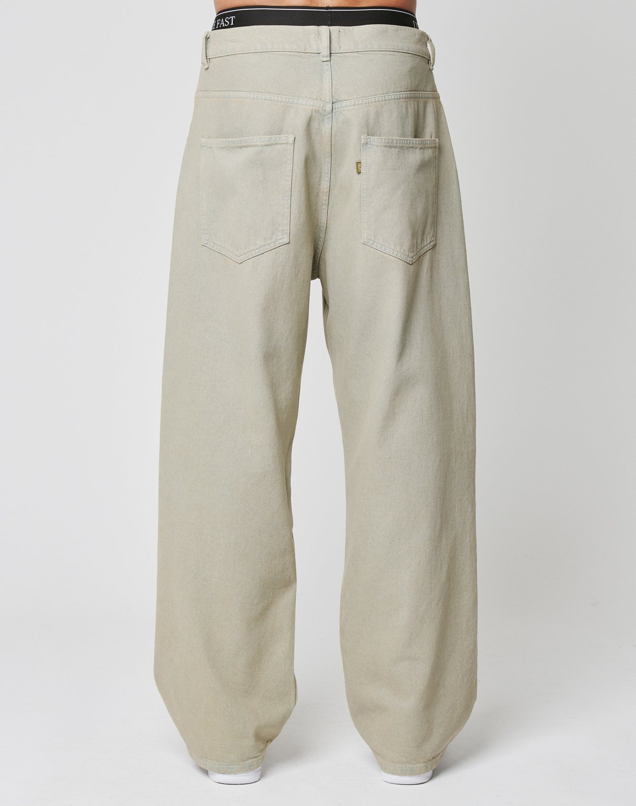 A person wearing loose-fitting beige LFDY Baggy Denim jeans by LIVE FAST DIE YOUNG is shown from the back, featuring two large back pockets, a visible black waistband above the denim, and a small Flag-Label on a white surface.
