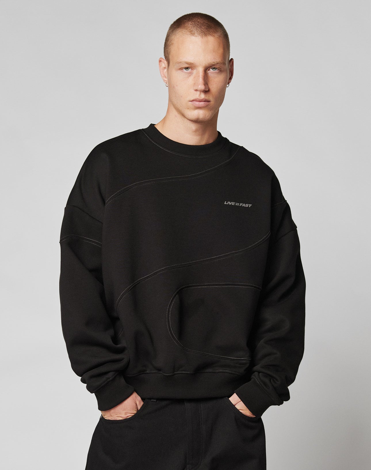 A person with short hair faces forward, wearing the oversize fit black LFDY Wavy Sweater by LIVE FAST DIE YOUNG—crafted from soft cotton with subtle curved seams and a bold LIFE IS FAST front print—paired with black pants against a plain light background.