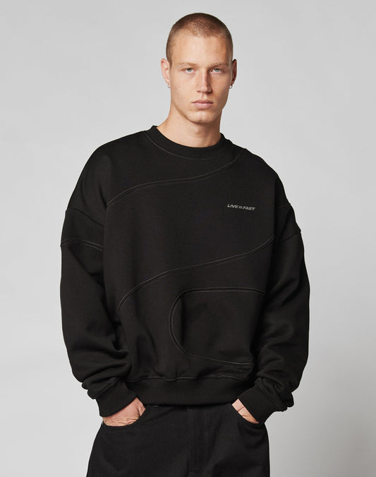 A person with short hair faces forward, wearing the oversize fit black LFDY Wavy Sweater by LIVE FAST DIE YOUNG—crafted from soft cotton with subtle curved seams and a bold LIFE IS FAST front print—paired with black pants against a plain light background.