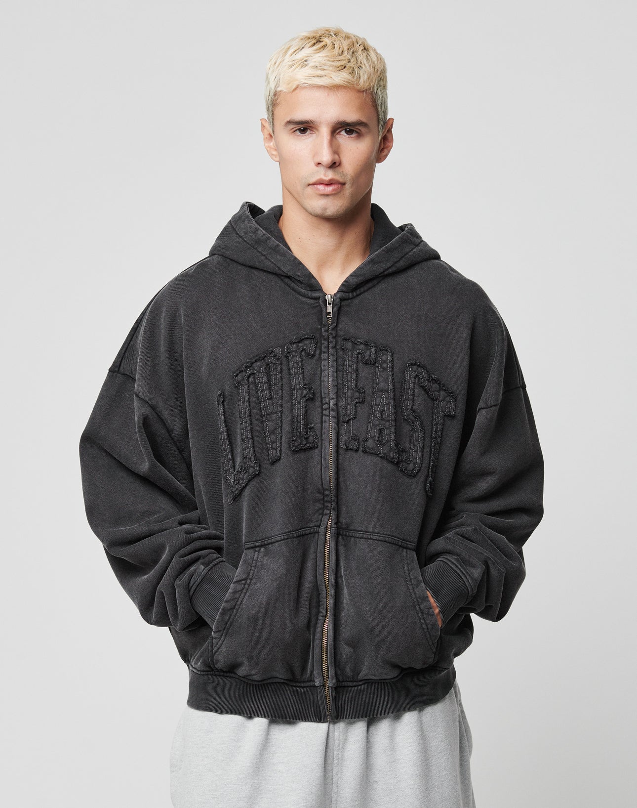 A young man with short blond hair wears the “Ripped College Hooded Zip” by LIVE FAST DIE YOUNG in Oversize Fit and gray sweatpants, standing against a plain light background.