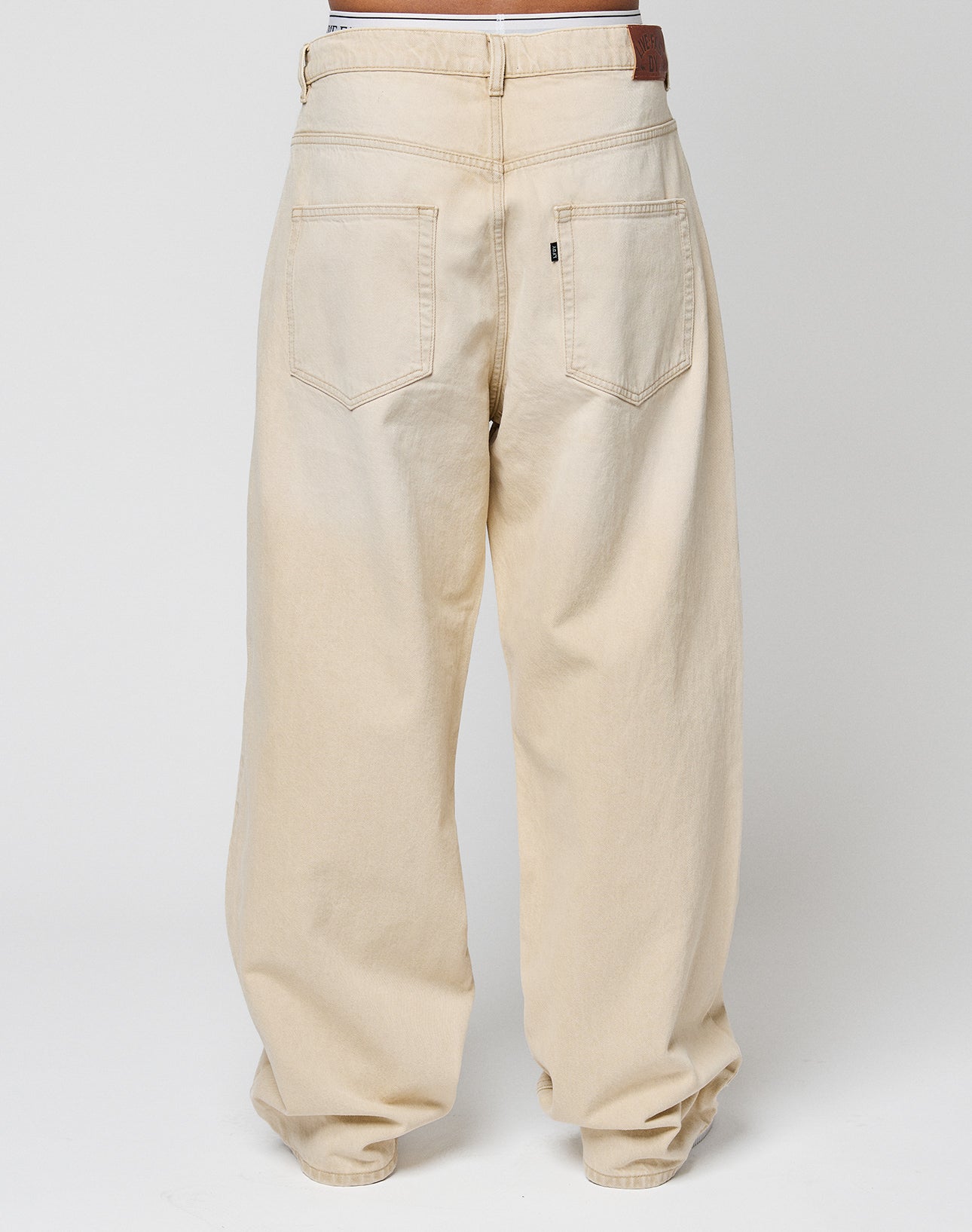 A person is shown from the back wearing LIVE FAST DIE YOUNGs LFDY Baggy Denim in light beige with a baggy fit and wide straight leg. Made from 100% Baumwolle, these jeans have two large back pockets and a brown leather patch on the waistband.