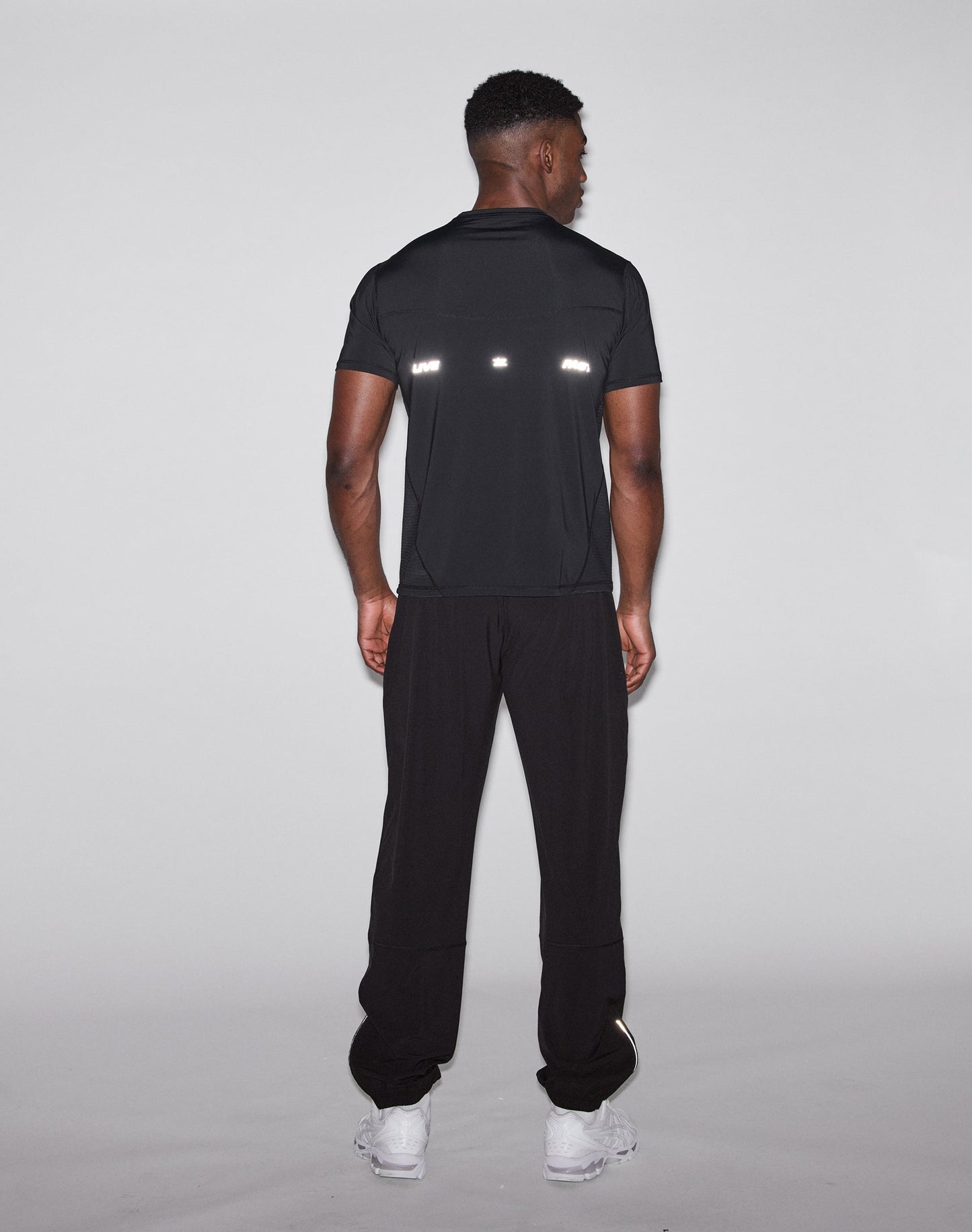 A man faces away from the camera wearing a LIVE FAST DIE YOUNG Live Fast Performance Tee, slim fit, black with reflective front and back print, paired with black pants and white sneakers against a plain light background.