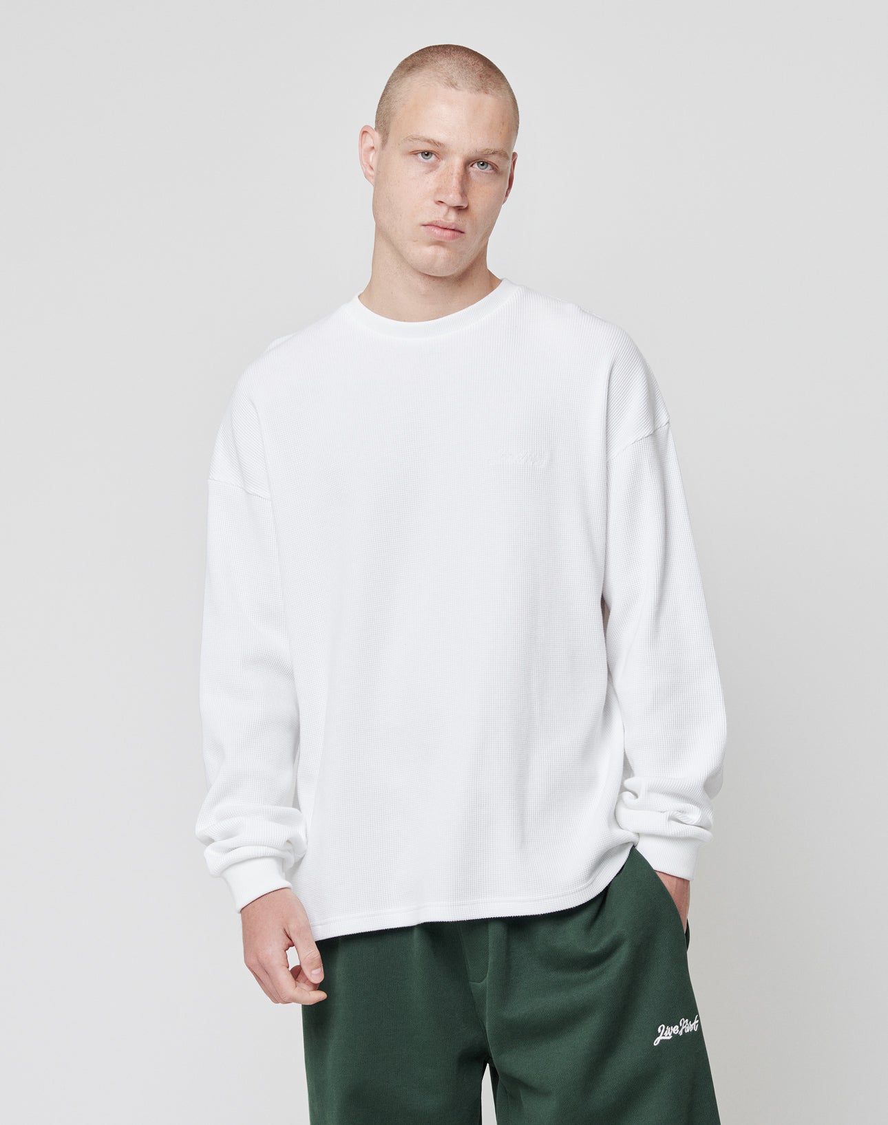 A person with short hair wears the LIVE FAST DIE YOUNG Basic Script Waffle Longsleeve in plain white and dark green pants, standing against a light gray background and looking at the camera with a neutral expression.