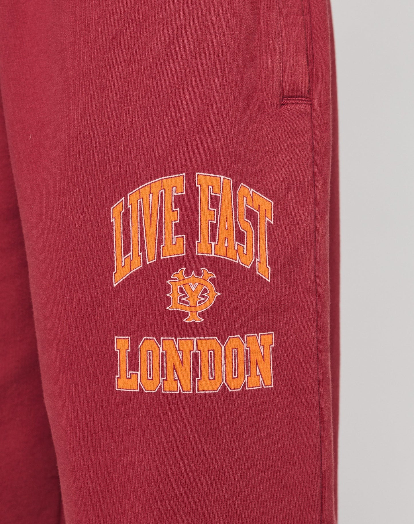 Close-up of the Live Fast London Wide Jogger by LIVE FAST DIE YOUNG: maroon wide-fit sweatpants with bold yellow and orange LIVE FAST LONDON text and emblem on the left thigh. Made from soft cotton with an elastic waist for comfort.