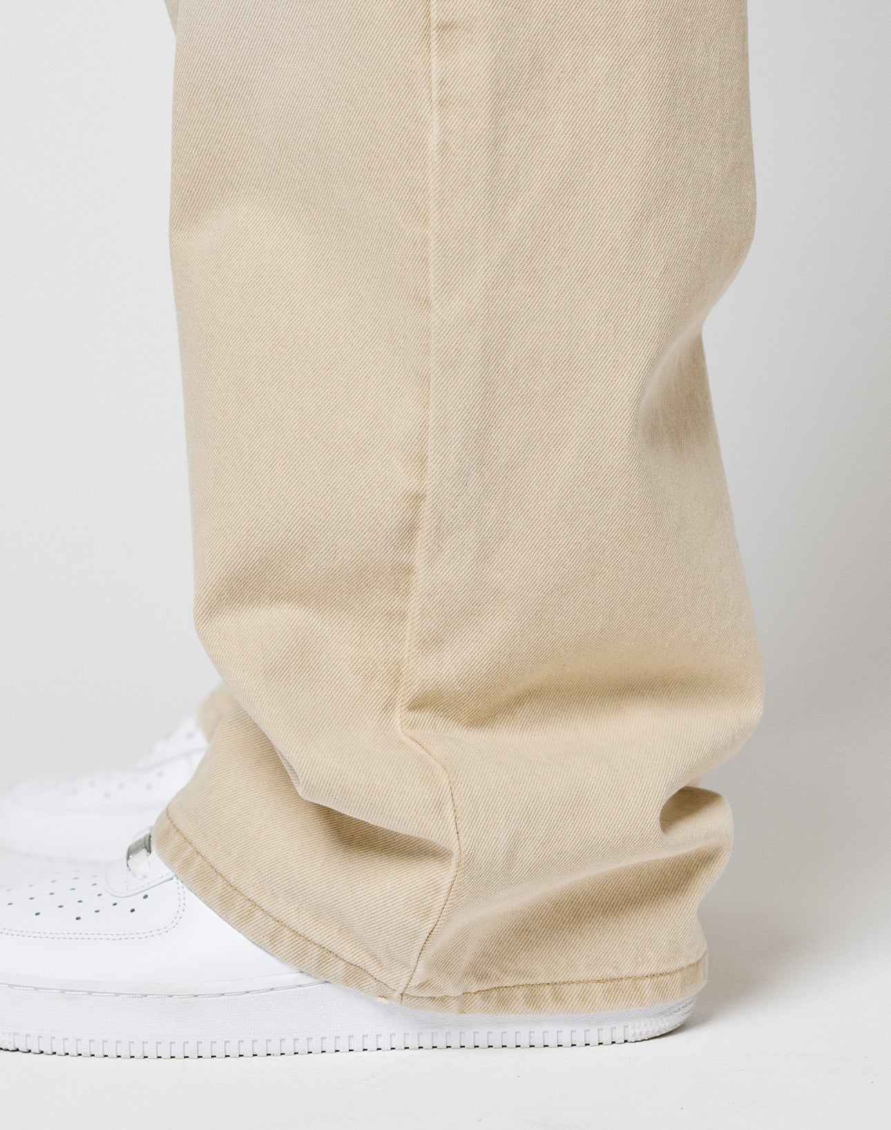 A close-up side view shows someone in LIVE FAST DIE YOUNG’s LFDY Baggy Denim—light beige, baggy fit with a wide straight leg and 100% cotton—slightly covering white sneakers against a plain, light background.