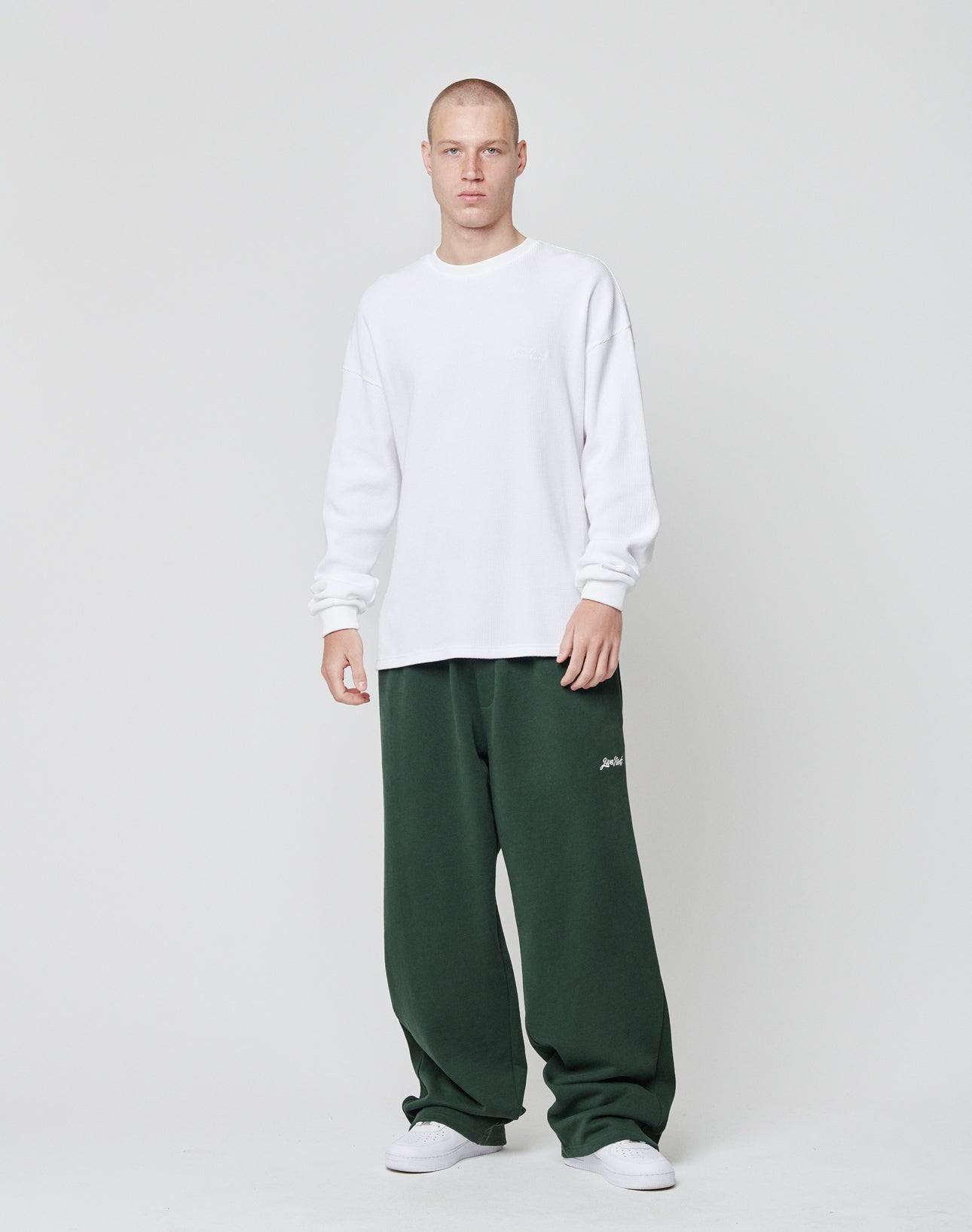A person stands against a plain white background, wearing the LIVE FAST DIE YOUNG Basic Script Waffle Longsleeve, paired with baggy dark green sweatpants and white sneakers for a casual, relaxed vibe.