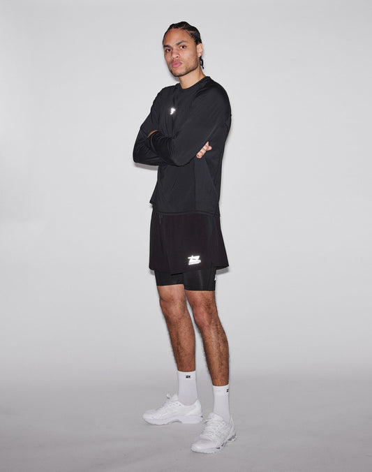 A person stands with arms crossed, wearing a black Regular Fit long-sleeve shirt, LIVE FAST DIE YOUNG Live Fast Performance Running Shorts in black with integrated tights, white socks, and white sneakers against a light-gray background.