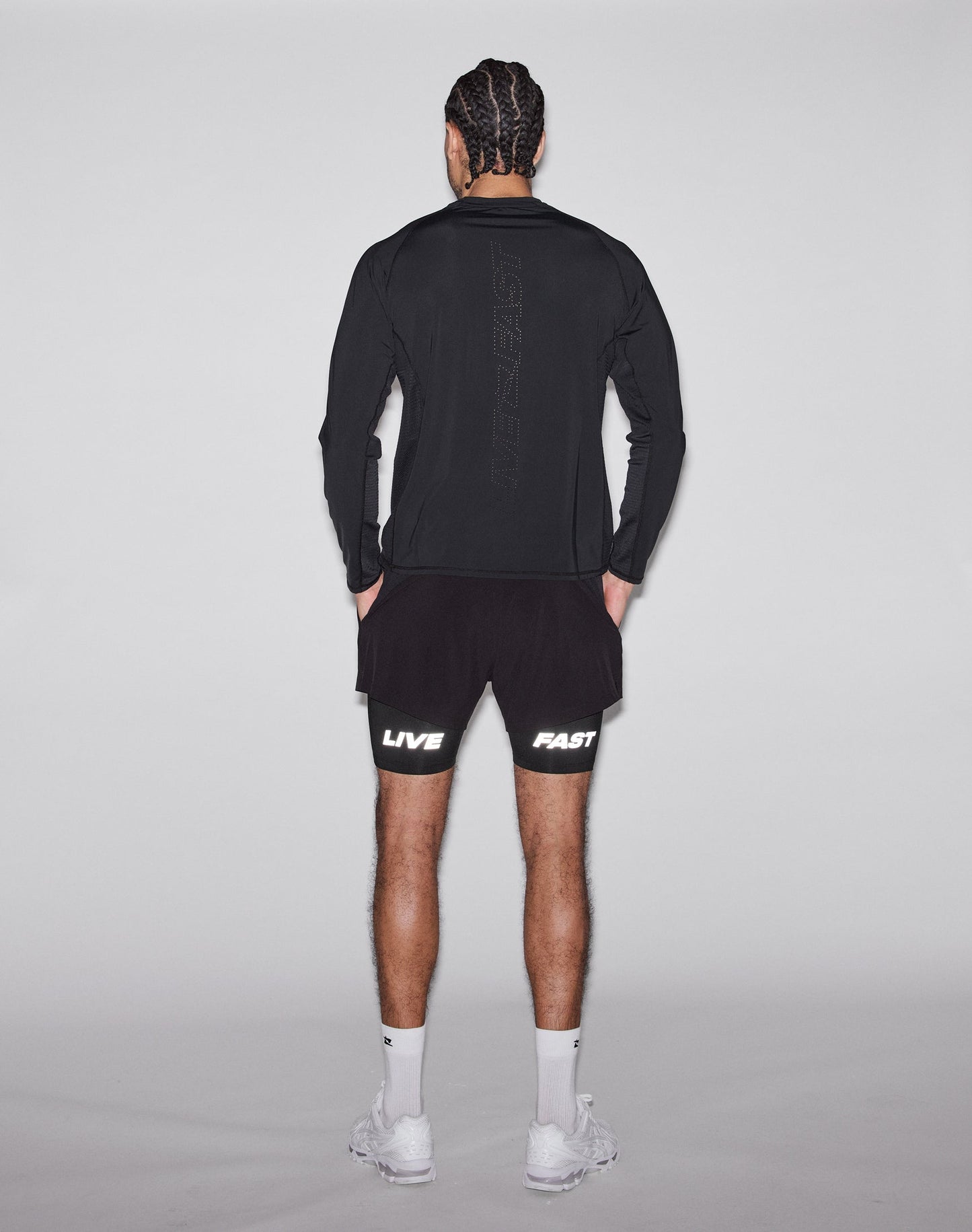A man with braided hair, white socks, and sneakers faces away wearing LIVE FAST DIE YOUNGs black Live Fast Performance Running Shorts (Regular Fit) featuring LIVE on the left leg and FAST on the right, plus a black long-sleeve shirt.