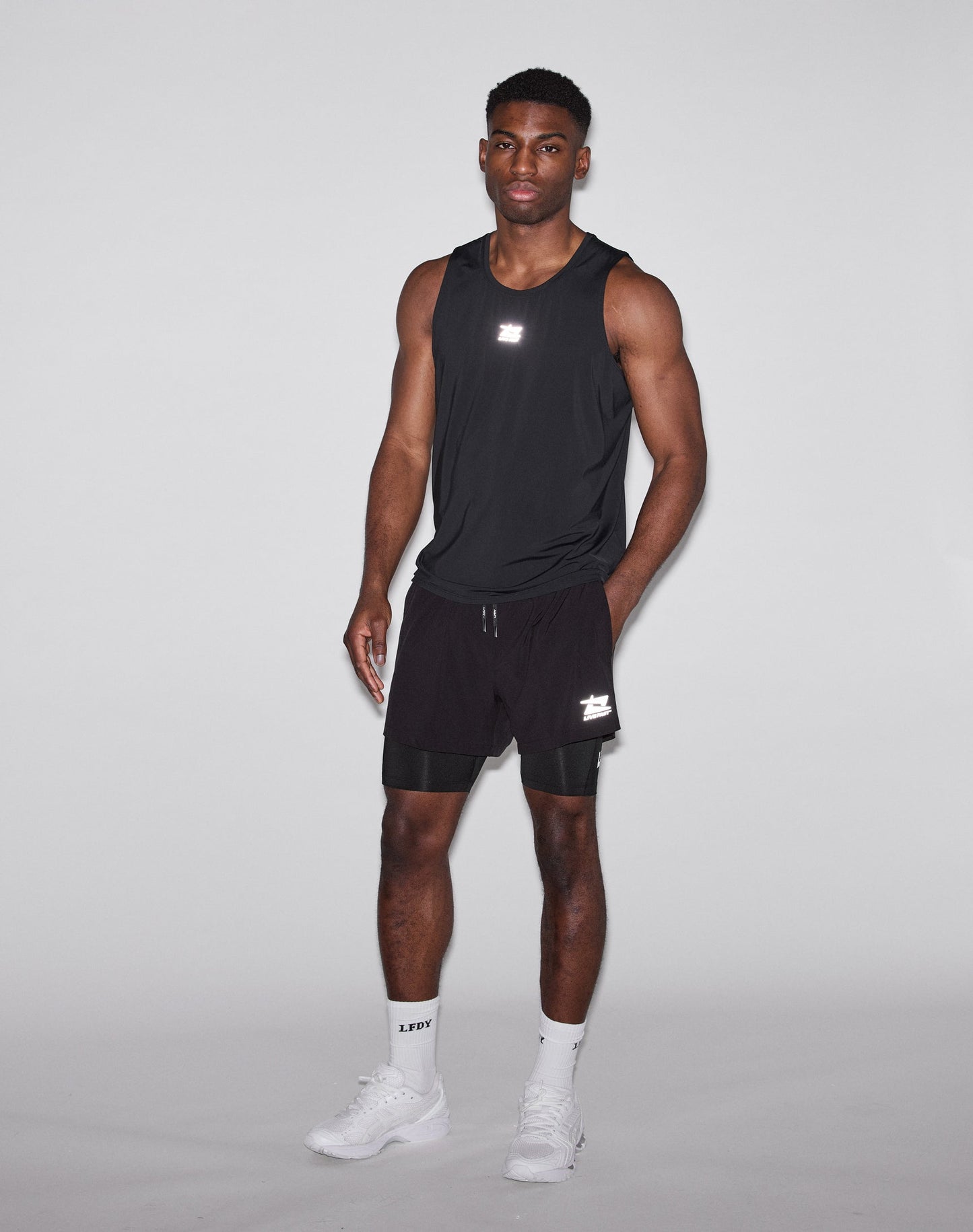 A man with short hair wears the LIVE FAST DIE YOUNG Live Fast Performance Tank, a slim fit sleeveless black athletic shirt in Nylon Spandex. LIVE FAST and a small reflective logo show in white on the back. He stands before a plain light background.