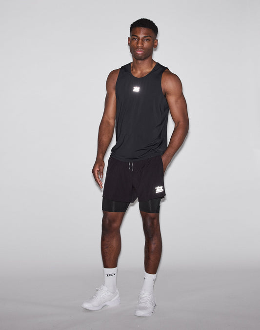 A man in a black LIVE FAST DIE YOUNG Live Fast Performance Tank, black shorts, white crew socks, and white sneakers stands against a white background with a serious expression and his left hand in his pocket.