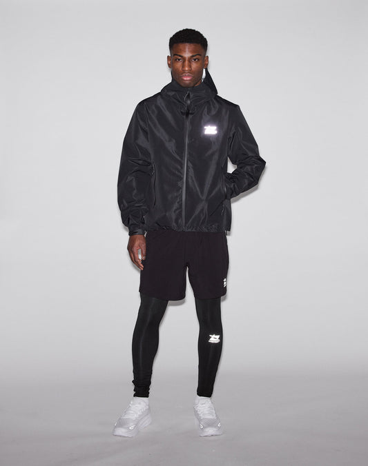 A man stands against a plain background wearing the LIVE FAST DIE YOUNG Live Fast Performance Rain Jacket with wasserdichte Nähte, black shorts and leggings, white sneakers, and reflective logos. His hands are at his sides.